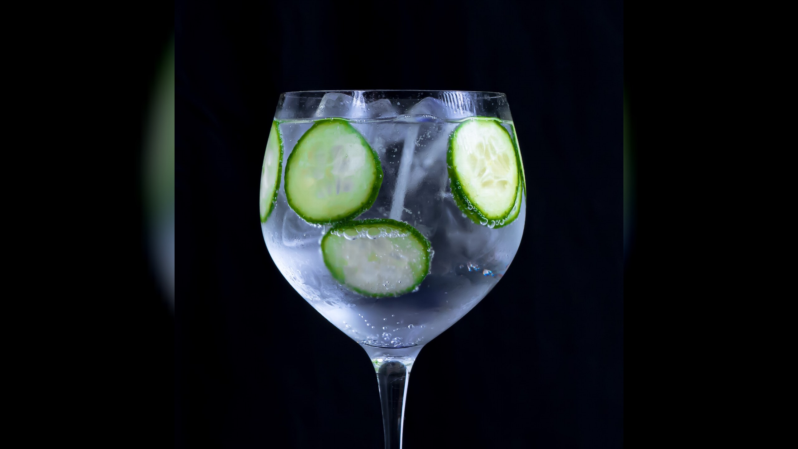 Gin Tonic im Copa-Glas mit Gurkenscheiben und Eis vor schwarzem Hintergrund